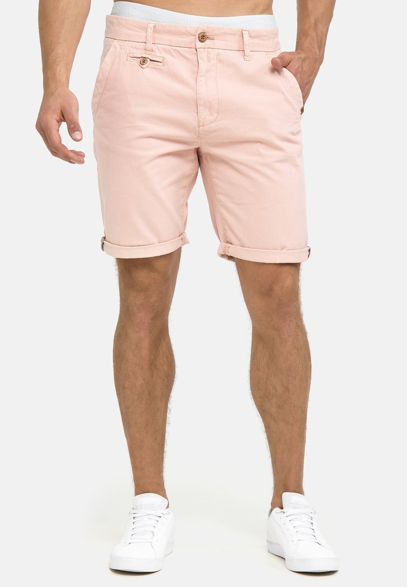 Shorts en coton rose clair avec des poignets retroussés, deux poches avant et une fermeture à bouton. Portés avec des baskets blanches sur un fond uni.