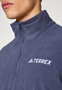 Granatowa kurtka polarowa z wysokim kołnierzem, zamkiem błyskawicznym i białym haftowanym logo „TERREX” na lewej piersi. Miękka faktura, dopasowany krój.