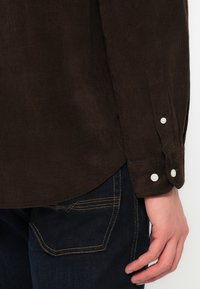Brun kordfløyelskjorte med hvite knapper, med teksturert stoff. Kombinert med mørkeblå jeans med kontrasterende gult søm.