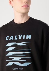 Sweatshirt preto com decote redondo, com texto gráfico em azul claro e elementos de design impressos na frente.