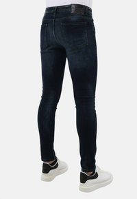 Dunne donkerblauwe jeans met een vervaagde textuur, voorzien van achterzakken en een zwart logo op de tailleband, gedragen met wit en zwarte sneakers.