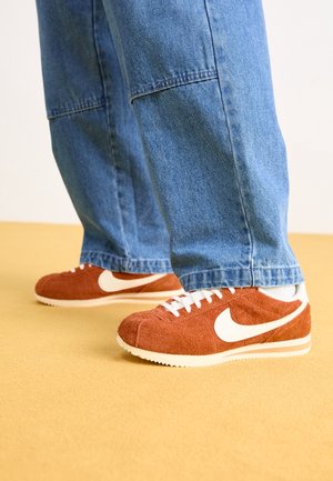 Osoba nosząca niebieskie spodnie dżinsowe i rdzawo-brązowe buty sportowe Nike z białymi logo Swoosh na żółtej podłodze.