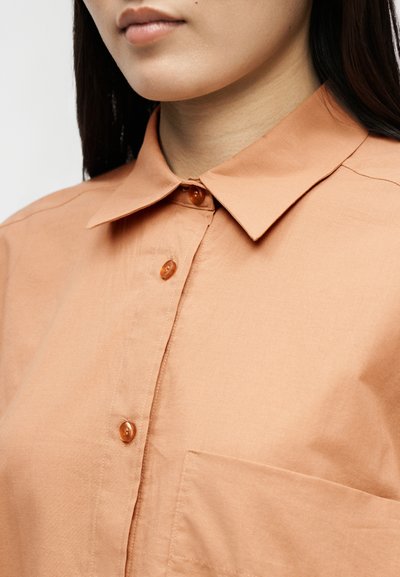 Žena oblečená v svetlohnedej košeli na gombíky s golierom, tromi viditeľnými gombíkmi a náprsným vreckom, zobrazená od brady po hornú časť trupu.
