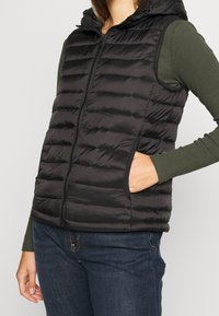 Svart isolerad väst med dragkedja framtill, pösiga paneler, huva och sidofickor, bärs över en grön långärmad tröja och mörka jeans.