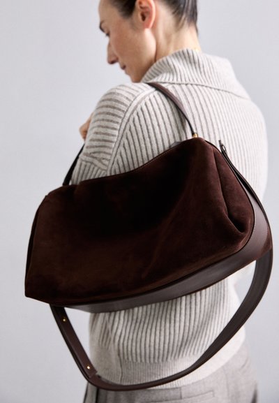 Sac hobo en suède marron avec une bandoulière en cuir courbée, doté d'une fermeture éclair et d'une finition lisse. Le sac a une forme spacieuse et structurée.