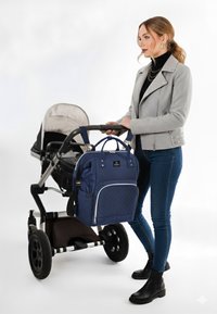 Blå kviltad ryggsäck med justerbara remmar, bärs av en person bredvid en barnvagn med stora svarta hjul och en grå barnstol.