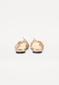 Gold strappy sandals with a flat sole, featuring a textured upper and decorative accents, open back, and logo on the heel.