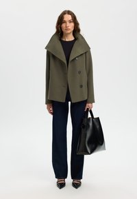 Olive green wool-blend coat with a high collar and four black buttons, worn over a dark top, paired with dark jeans and black tote bag.