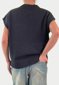 Maglione nero a maglia con maniche corte, colletto e orlo a coste. Indossato con pantaloni di jeans azzurri, mani nelle tasche.