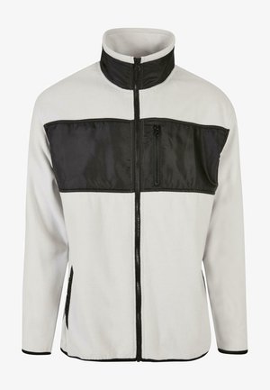 Világosszürke fleece zipzáras kabát, fekete nylonzsebbel a mellrészen, magas nyakkal, oldalsó zsebekkel és elöl zipzárral.