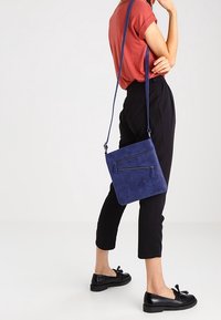 Malá tmavě modrá semišová kabelka přes rameno se dvěma předními zipy, dlouhým nastavitelným popruhem a obdélníkovým tvarem. Hladká textura.