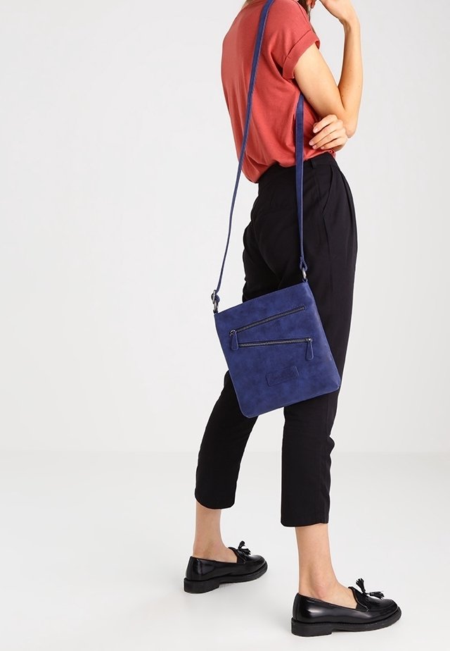 Malá tmavě modrá semišová kabelka přes rameno se dvěma předními zipy, dlouhým nastavitelným popruhem a obdélníkovým tvarem. Hladká textura.