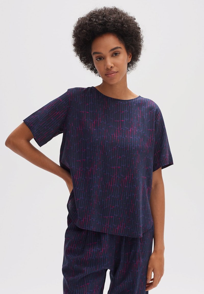Donna con capelli ricci che indossa un abbigliamento coordinato blu navy composto da top e pantaloni con un motivo geometrico rosso, in piedi contro uno sfondo chiaro e uniforme.
