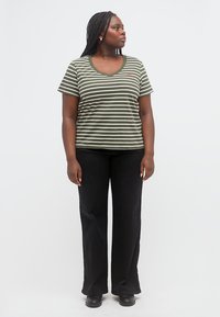 Γυναίκα με πλεγμένα μαλλιά που φοράει πράσινη και λευκή ριγωτή μπλούζα με V-λαιμόκοψη, μαύρο παντελόνι και μαύρα παπούτσια, στέκεται και κοιτά προς το πλάι.