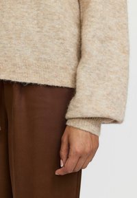 Bež prugasti pulover z mehkimi strukturami in rebrastimi manšetami, oblečen preko rjavih hlač. Vidna roka z nevtralnim lakom za nohte.