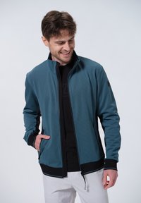 Giacca leggera con zip in teal, caratterizzata da un motivo testurizzato, colletto alto e dettagli neri. Due tasche laterali e branding minimale visibile.