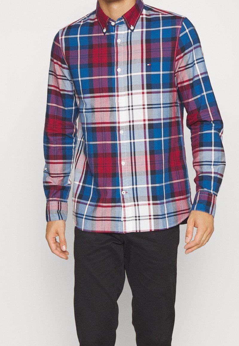 Homme portant une chemise à carreaux boutonnée à manches longues rouge, bleu, blanc et noir avec un pantalon noir sur un fond clair uni.