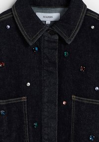 Camisa vaquera en azul oscuro con cuello clásico, dos bolsillos delanteros y adornos de joyas multicolores. Costuras en dorado que resaltan.