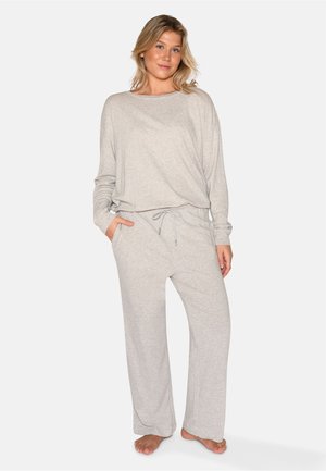 Femme portant un haut gris clair à manches longues et un pantalon ample assorti, debout pieds nus, une main dans la poche, devant un fond uni.