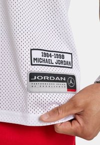 Hvid mesh jersey med sorte og grå etiketter; den ene læser "1984-1998 MICHAEL JORDAN," og den anden "JORDAN PERFORMANCE BRAND."