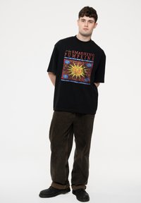 Black oversized t-shirt featuring a graphic design with a yellow sun and multicolored accents. Paired with brown corduroy pants and black shoes.