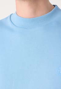 Gros plan sur une personne portant un pull ras du cou bleu clair avec un petit logo circulaire en relief sur la poitrine.