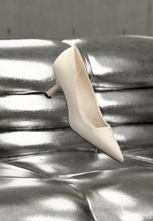 Zapato de tacón alto de punta estrecha en material blanco brillante con un tacón delgado. El diseño presenta un acento negro en el tacón y una textura lisa.