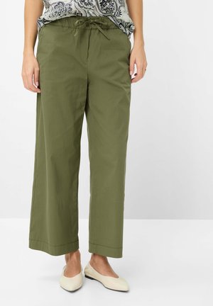 Frau trägt olivgrüne, weit geschnittene Hosen mit Kordelzug an der Taille und cremefarbene flache Schuhe, stehend vor einem neutralen Hintergrund.