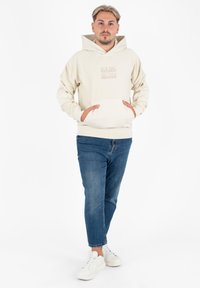 Beżowy polarowy hoodie z przednią kieszenią, kapturem ze sznurkiem i subtelny logo. Noszony z niebieskimi dżinsami i białymi trampkami.