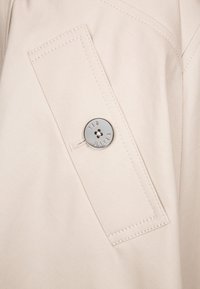 Zbliżenie na beżową tkaninę z kieszonką, w której znajduje się duży srebrny guzik z wygrawerowanym napisem "TED BAKER." Gładka tekstura z widocznymi detalami nici.