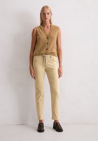 Vrouw met steil blond haar draagt een mouwloze bruine vest met knopen, beige broek met aansnoerkoord en donkerbruine instappers, staande tegen een witte muur.