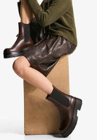 Botine maro din piele cu panouri elastice pe lateral, talpă groasă din cauciuc negru și vârf rotunjit, asortate cu o fustă maro texturată.