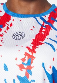Bílý sportovní triko s modrými a červenými abstraktními vzory, síťovina, kulatý výstřih, s kruhovým logem uprostřed.