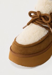 Bottines en suède brun avec une douce doublure en polaire crème, une semelle épaisse, et des lacets en corde brun avec des embouts en métal. Forme arrondie et finition texturée.