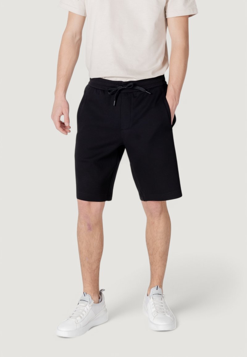 Pantaloni corti neri in cotone con cintura elastica, coulisse e tasche laterali, abbinati a sneakers bianche e una maglietta di colore chiaro.