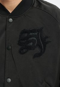 Svart bomberjakke med et teksturert, brodert logo på fronten. Glatt stoff med trykknapplukking og ribbestrikket krage.