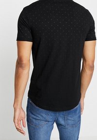 Camiseta negra de manga corta con un patrón sutil, cuello redondo y un dobladillo ligeramente curvado, combinada con jeans azules.