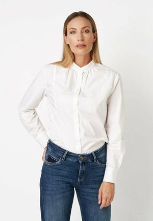 Woman with long blonde hair wearing a white buttoned shirt and blue jeans, standing against a plain light background, facing forward.
