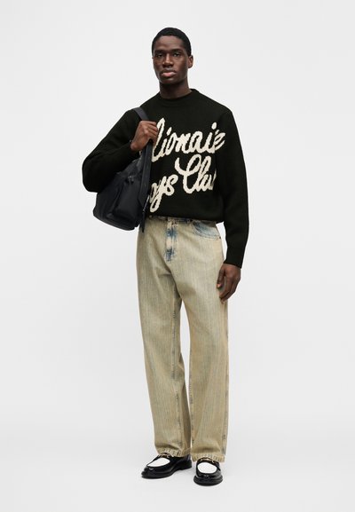 Μαύρο πλεκτό πουλόβερ με λευκό κείμενο, ανοιχτό μπλε ψηλόμεσο τζιν με ξεθωριασμένο σχέδιο και μαύρα μοκασίνια. Κρατάει μια μαύρη τσάντα ώμου.