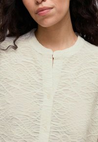 Light cream button-up blouse with a textured wave pattern, featuring a round neckline and subtle sheen on a sheer fabric.