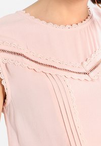 Light pink blouse with lace trim, featuring a round neckline and pleated design. Soft fabric with a smooth texture and decorative accents.