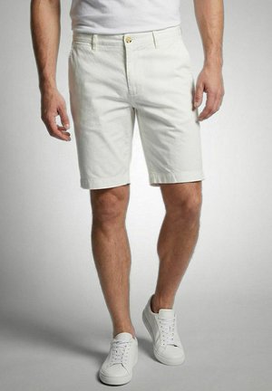 Man met witte knielange shorts, witte sneakers en een wit T-shirt, staand met licht gespreide benen op een grijze achtergrond.