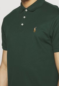 Tmavě zelené pánské polo tričko vyrobené z bavlny, s tříknoflíkovou légou, krátkými rukávy a malým vyšívaným logem na levé hrudi.