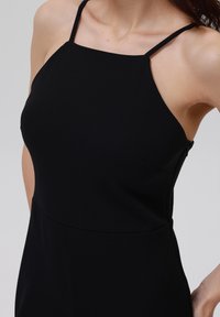 Schwarzes ärmelloses Kleid mit einem hohen Ausschnitt, dünnen Schulterträgern, strukturiertem Stoff und einer figurnahen Silhouette mit einem unauffälligen Reißverschluss an der Seite.