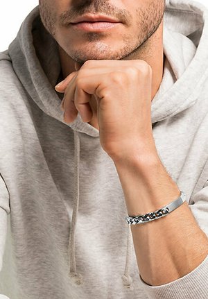 Silberne Kettenarmband mit einem flachen Band und einem strukturierten Glieddesign, getragen an einem Handgelenk, vor einem hellgrauen Hoodie-Hintergrund.