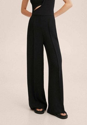 Frau in weiten schwarzen Hosen, einem ärmellosen schwarzen Top mit seitlichem Cut-out und schwarzen Plateausandalen vor einem schlichten Hintergrund.