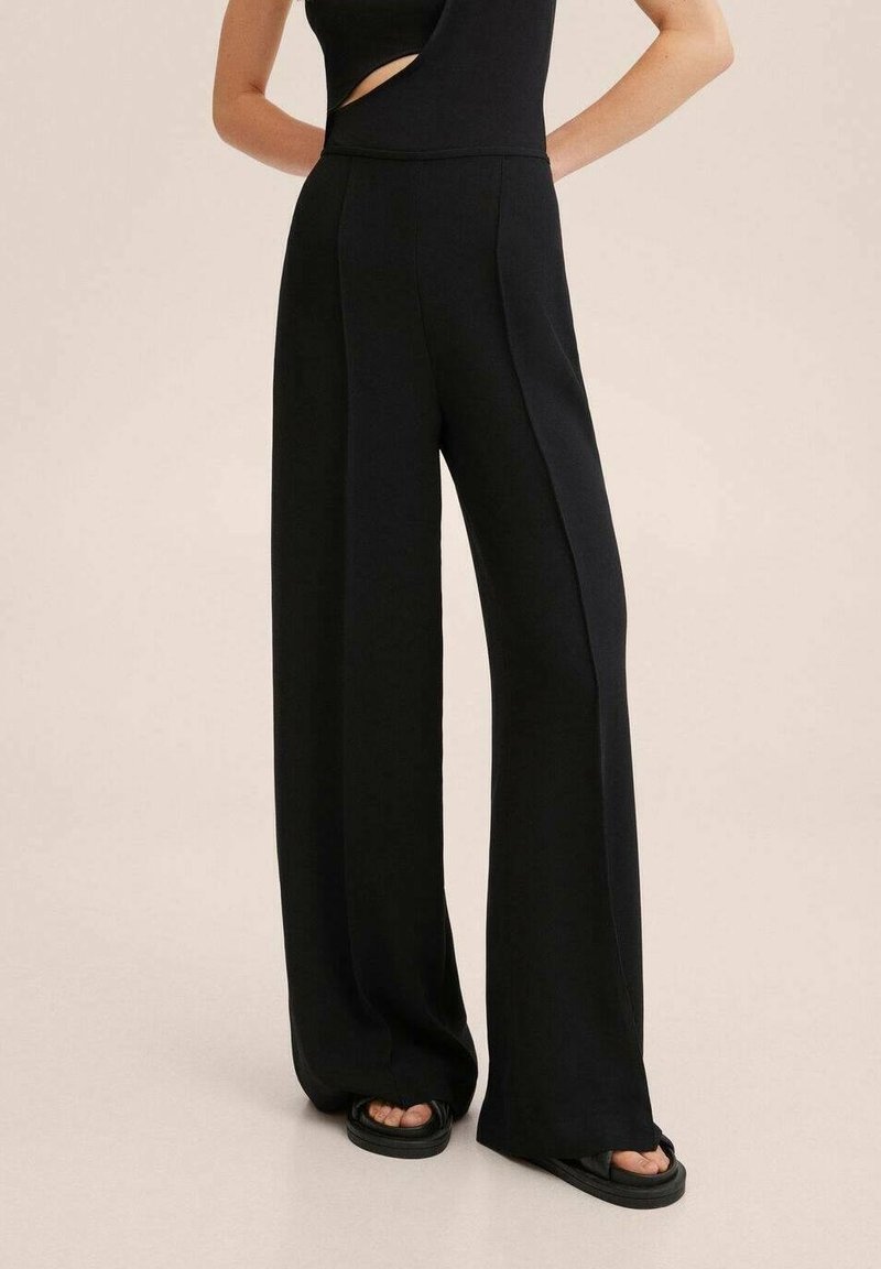 Femme portant un pantalon noir à jambes larges, un haut noir sans manches avec une découpe sur le côté et des sandales noires à plateforme, sur fond uni.