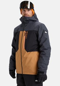 Mann, der eine braun-schwarze Winterjacke, schwarze Handschuhe, eine orangefarbene Mütze und Skibrille trägt, steht vor einem weißen Hintergrund.