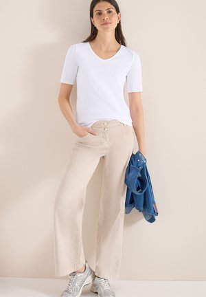 Femme debout, une main dans la poche, portant un haut blanc à manches courtes, un pantalon beige à jambes larges, des baskets argentées, tenant une veste en denim bleu.