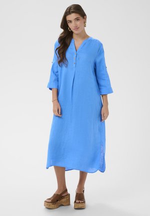 Žena s dugom smeđom kosom u lepršavoj plavoj midi haljini s gumbima i smeđim platformskim sandalama, stoji na jednobojnoj bijeloj pozadini.